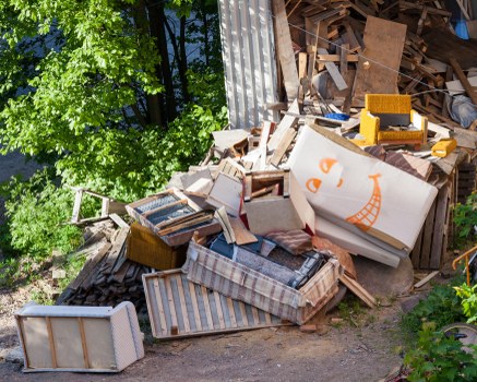 A large pile of discarded household furniture and wooden debris situated outdoors on a concrete surface, next to a corrugated metal fencing on the right. The pile includes a variety of items such as a striped upholstered armchair, wooden cabinets, and broken furniture pieces, with some items stacked haphazardly and others leaning against the fence. The furniture surfaces show signs of wear, with chipped paint and scratched finishes, and some items are partially dismantled. Bright sunlight illuminates the scene, casting shadows across the clutter. Surrounding greenery, including a leafy tree on the left, indicates the pile is located in an outdoor area, potentially a driveway or garden space. The environment and the presence of mixed waste materials suggest this is an area where rubbish removal services by Commercial Waste Bayswater could be applicable for clearing unwanted household items and debris in the Bayswater locality near London.