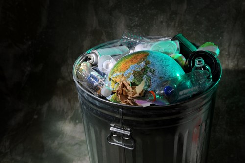 A metal waste bin with a hinged lid, filled with various pieces of discarded plastic bottles, caps, and a crumpled piece of brown paper, with a small globe of Earth resting among the debris. The bin is positioned outdoors on a dark, textured surface, likely on a pavement or driveway, with a dimly lit background that emphasizes the reflective surface of the bin and the colorful plastics. The assortment of plastics varies in clarity, with some bottles being transparent and others having colored tints, and the globe features blue, green, and brown tones representing continents and oceans. The scene captures a moment of rubbish accumulation, relevant to waste removal services such as those provided by Commercial Waste Bayswater, serving the Bayswater area, with visual emphasis on waste materials that require proper disposal.