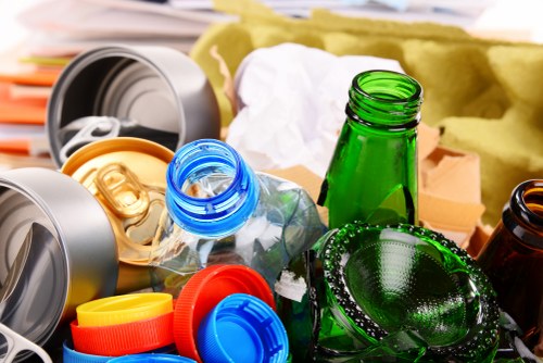 A close-up image showing a collection of discarded glass bottles, plastic containers, and aluminum cans piled together, with some bottles lying on their sides and others standing upright. The bottles are in various colours including green, clear, and amber, with some featuring labels or residual liquid. Surrounding the bottles are plastic caps in red, blue, and yellow, along with metallic lids and rings. In the background, part of a yellow paper or cardboard wrapping material and a glimpse of a wooden surface are visible, suggesting an outdoor residential or commercial setting suitable for rubbish removal services in Bayswater. The scene highlights common recyclable waste items that a waste removal company like Commercial Waste Bayswater might manage as part of their rubbish collection offerings.