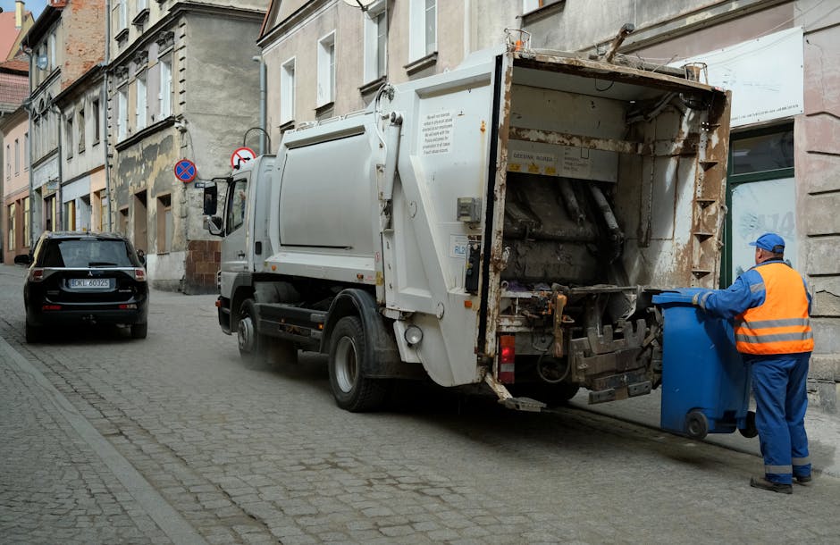 A small, aged blue utility truck parked alongside a street lined with trees and greenery, loaded with a large pile of mixed waste including flattened cardboard boxes, black plastic garbage bags, and v