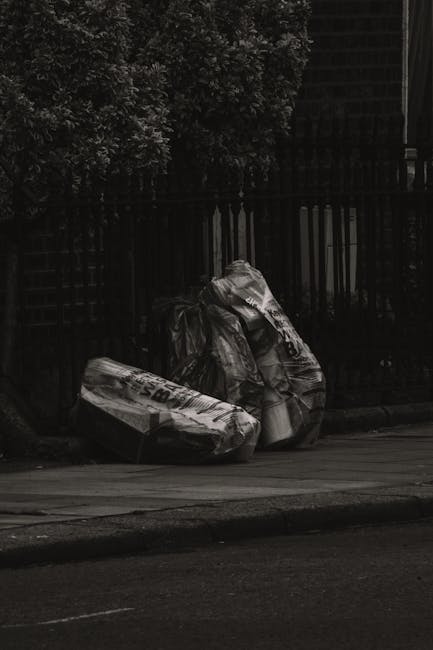 Two black garbage bags filled with waste materials, positioned on the edge of a paved street near a wooden fence and dense foliage. The bags appear to be made of plastic with a glossy finish, reflecti